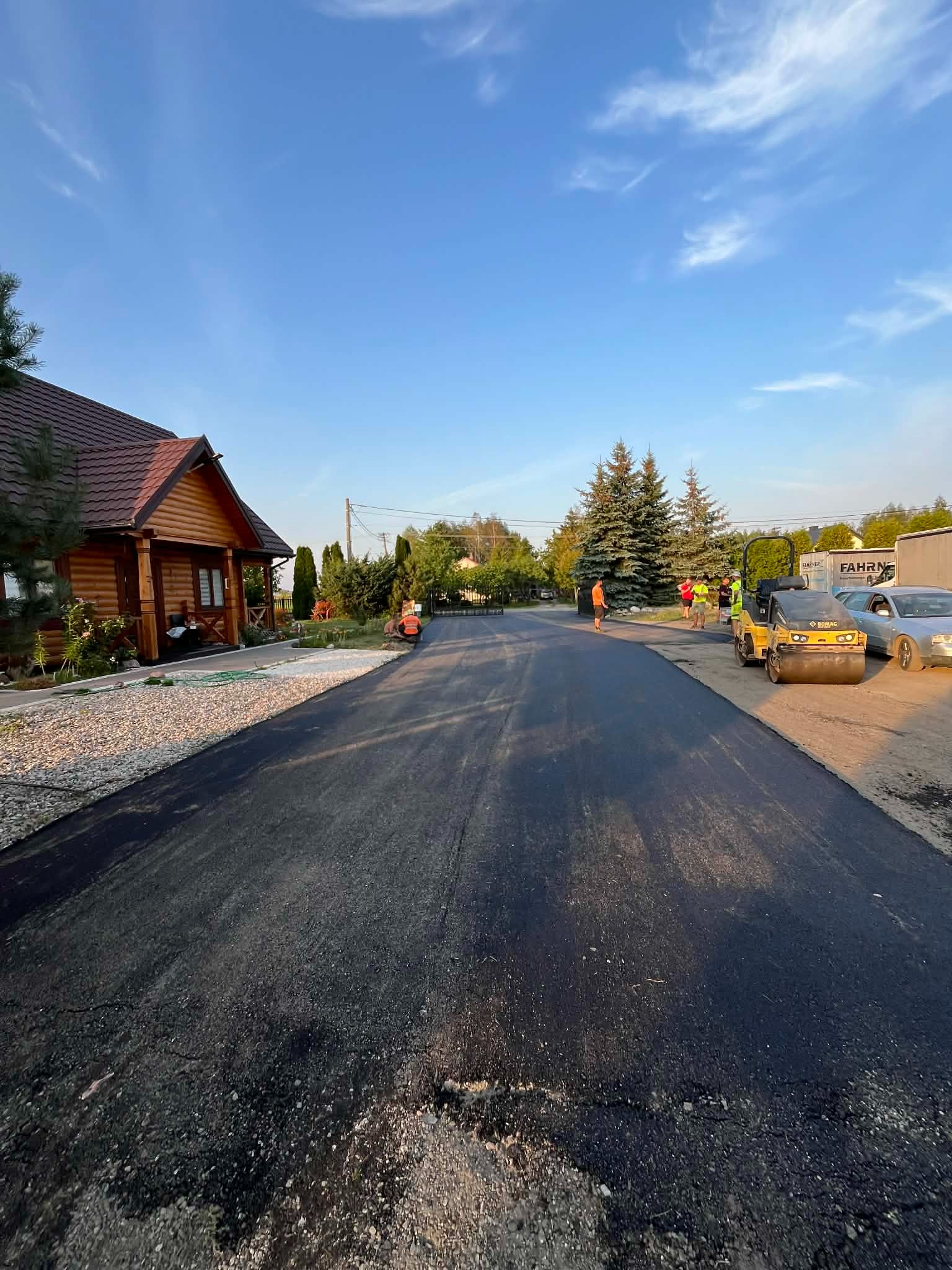 Świeżo położony asfalt na drodze dojazdowej, z widocznym walcem drogowym i ekipą pracowników w tle, obok drewnianego domu i zieleni.