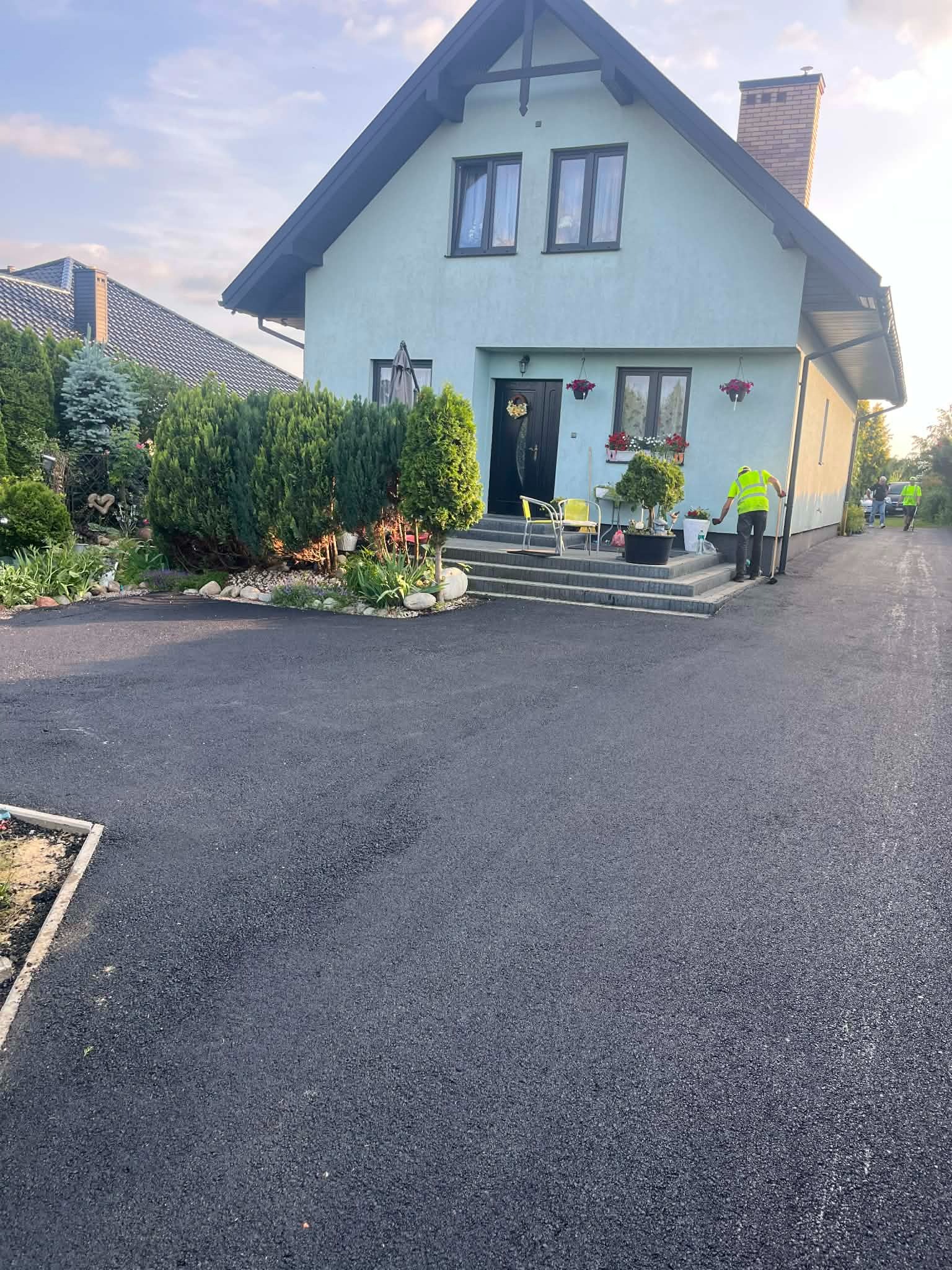 Nowa, czarna nawierzchnia asfaltowa przed domem jednorodzinnym. Pracownicy w odblaskowych kamizelkach. Świeżo położony asfalt, widoczne detale wykończenia.