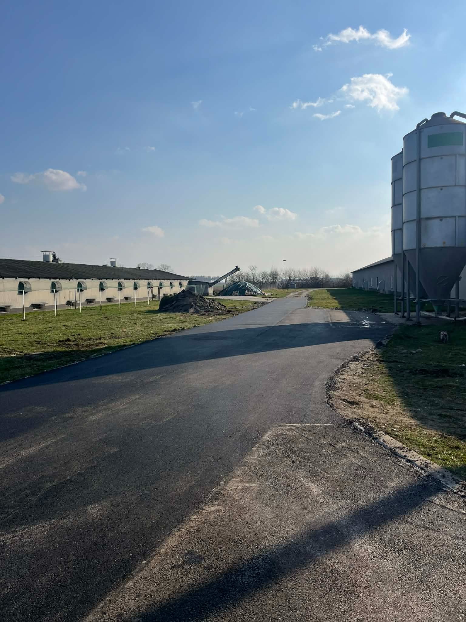 Nowa, asfaltowa droga dojazdowa na terenie gospodarstwa rolnego, z widocznymi silosami i budynkami gospodarczymi w tle, w słoneczny dzień.