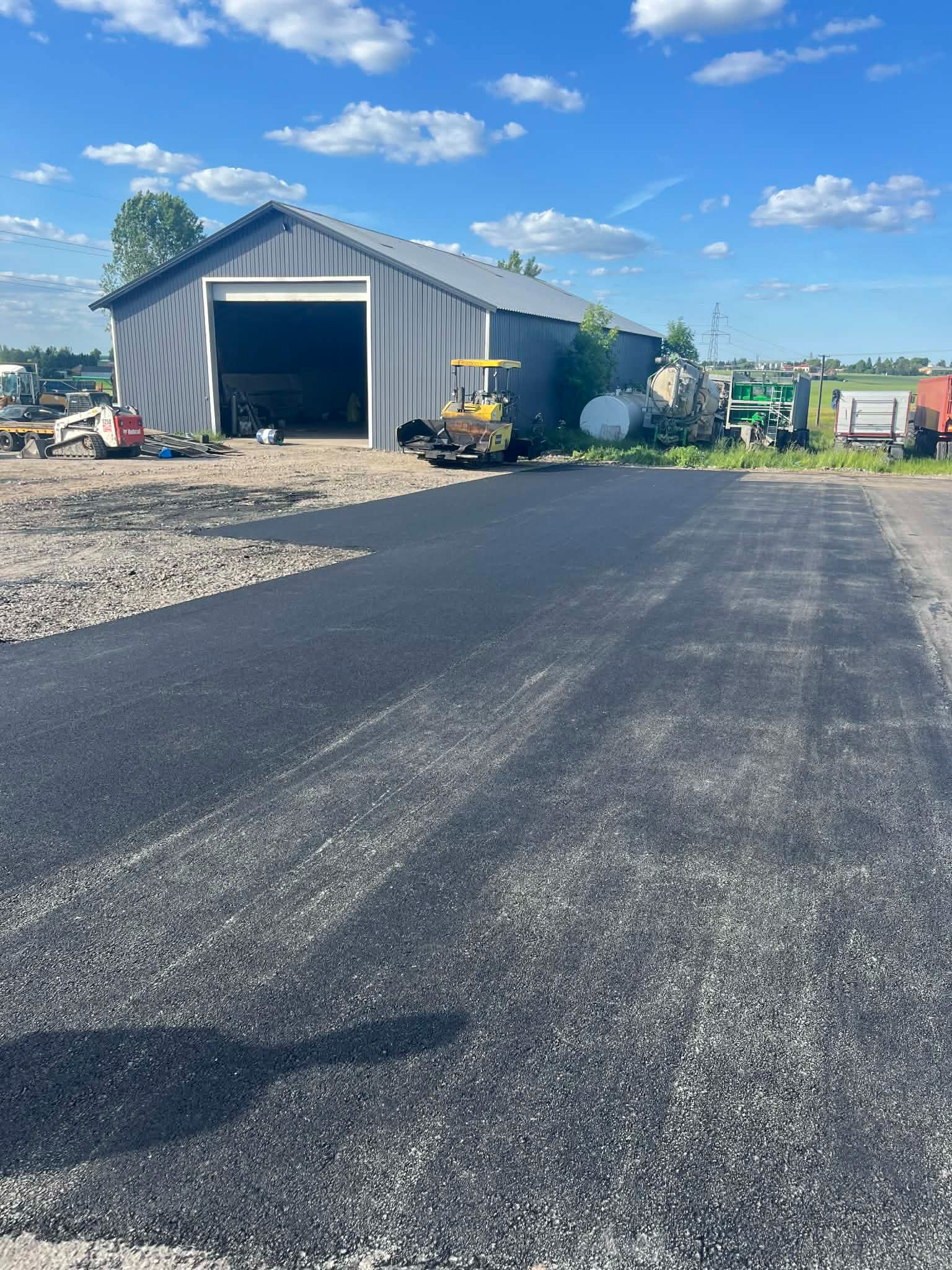 Świeżo ułożona, czarna nawierzchnia asfaltowa na placu, z budynkiem gospodarczym w tle i sprzętem budowlanym. Słoneczny dzień, błękitne niebo z chmurami.