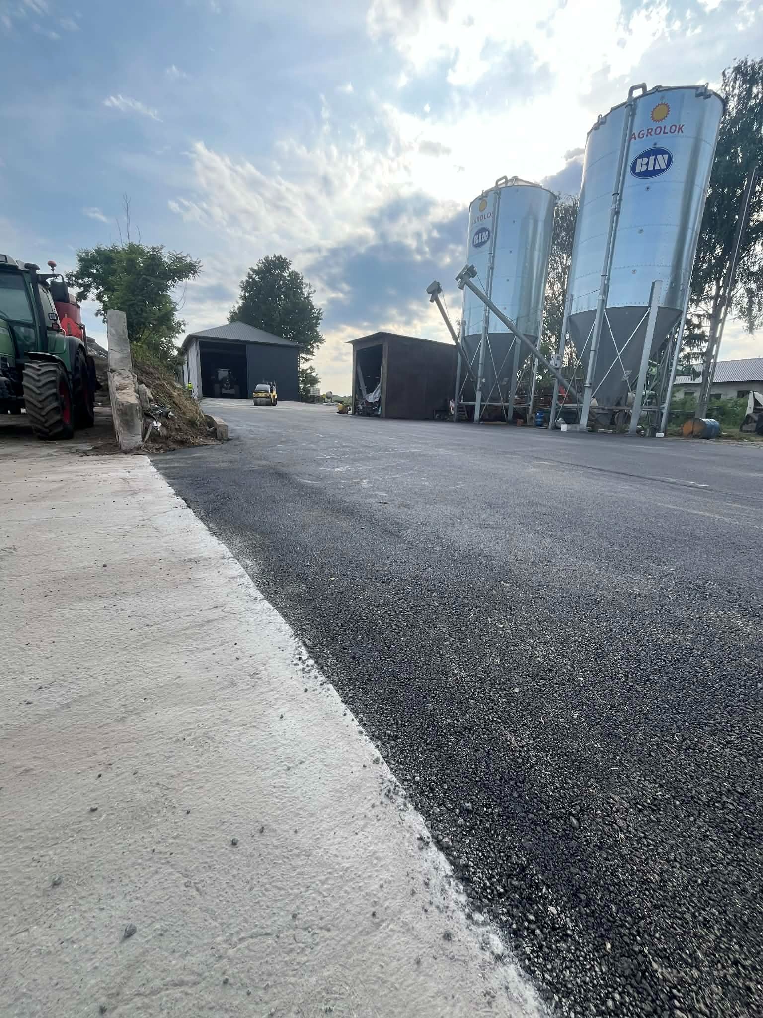 Nowa, czarna nawierzchnia asfaltowa z betonowym obrzeżem, maszyny rolnicze w tle i zbiorniki Agrolok. Świeżo położony asfalt na terenie gospodarstwa.