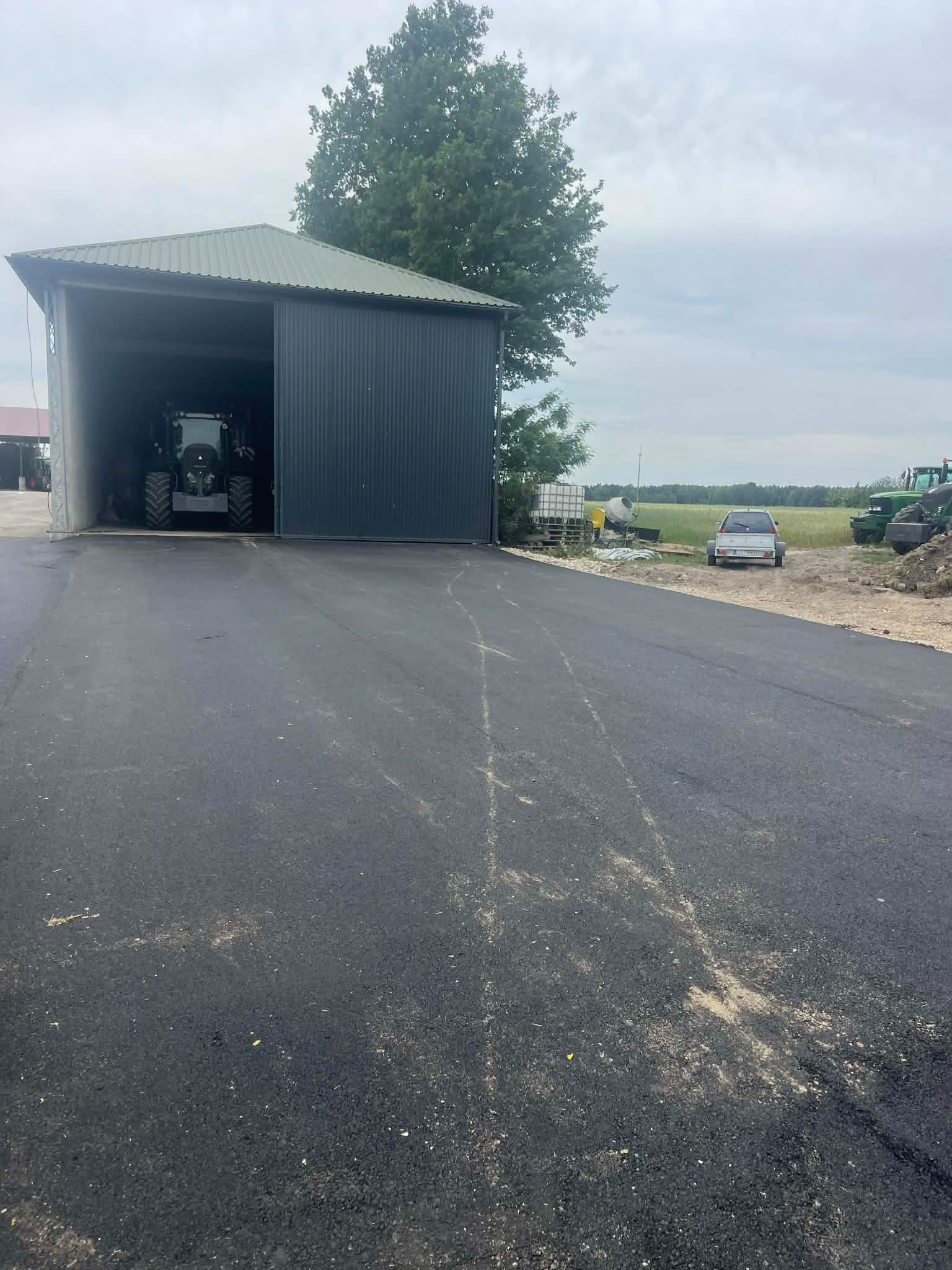 Nowa nawierzchnia asfaltowa w Warszawie, prowadząca do garażu z traktorem. W tle widoczne inne pojazdy i sprzęt budowlany na tle zieleni.