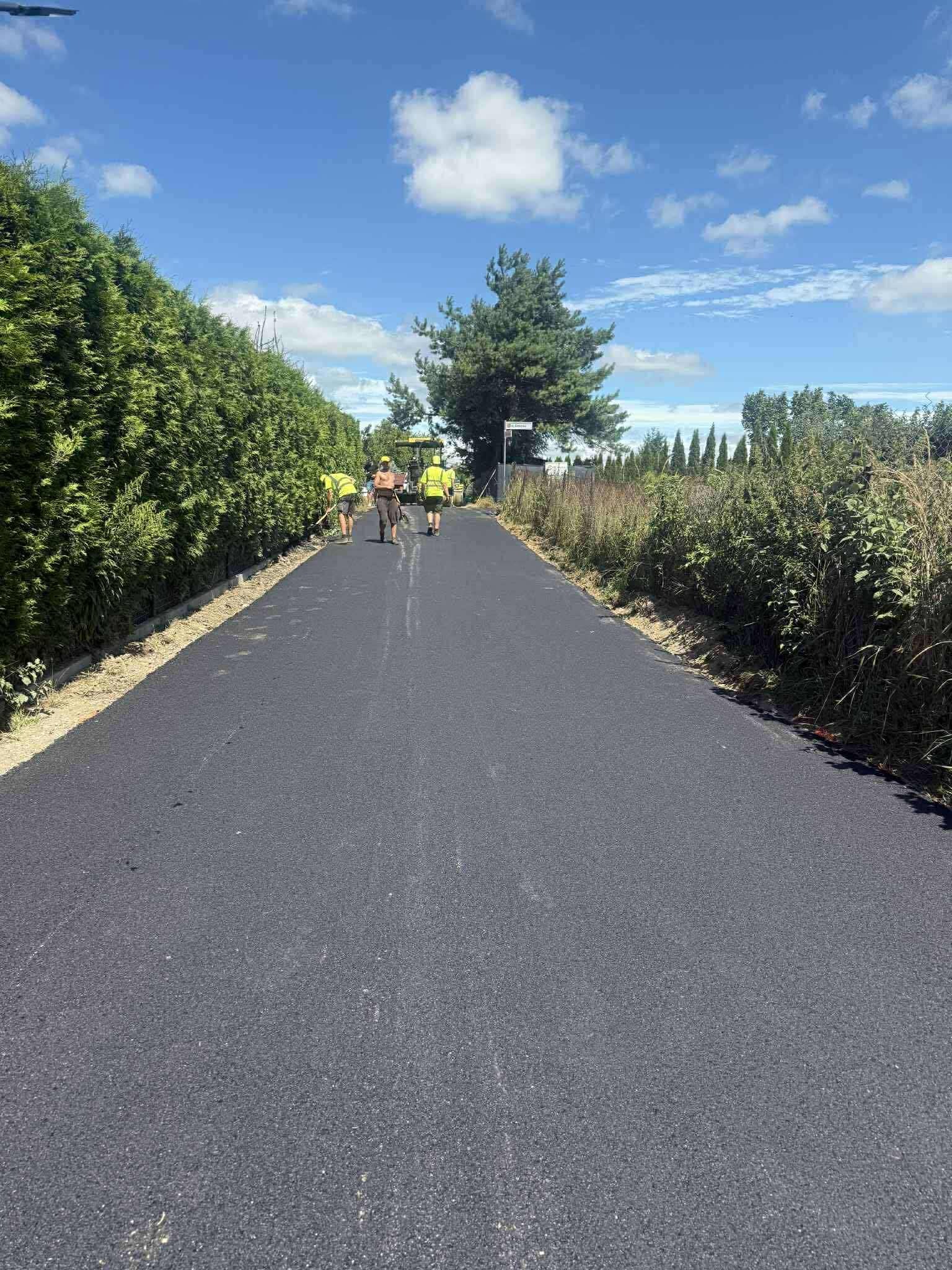 Świeżo położony asfalt na drodze osiedlowej, z pracownikami w odblaskowych kamizelkach i maszyną w tle, otoczonej zielenią w słoneczny dzień.