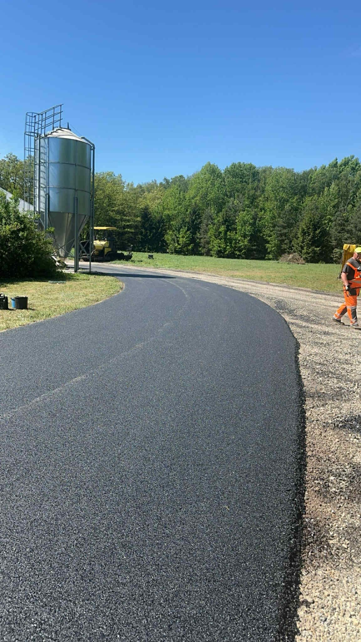 Nowo położona, czarna nawierzchnia asfaltowa drogi zakręcającej w prawo, z pracownikiem w pomarańczowym kombinezonie na poboczu i silosami w tle.
