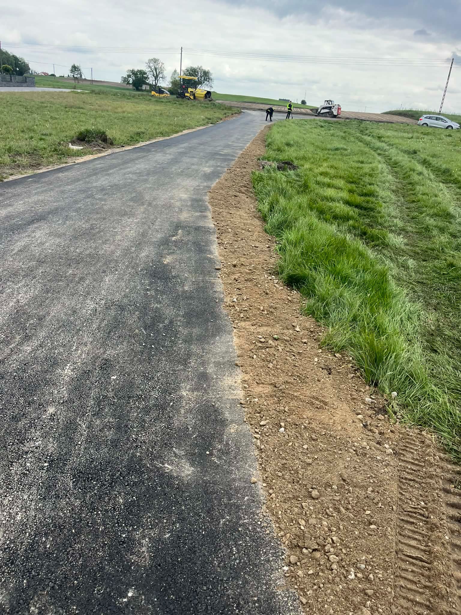 Nowa, asfaltowa droga na etapie budowy, z widocznymi maszynami budowlanymi i pracownikami w oddali, otoczona zielonym terenem. Świeżo położona nawierzchnia.