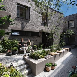 Michał Góral U3D - Nowoczesny dziedziniec z szarej cegły, gdzie para siedzi na ławce wśród zieleni. Kot śpi na murku, rower oparty o ścianę. Architektura minimalistyczna, rośliny w donicach.