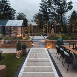 Michał Góral U3D - Stylowa aranżacja ogrodu z paleniskiem, szklarnią i jadalnią na zewnątrz. Uwagę przyciąga oświetlenie i dbałość o detale, tworząc przytulną atmosferę.