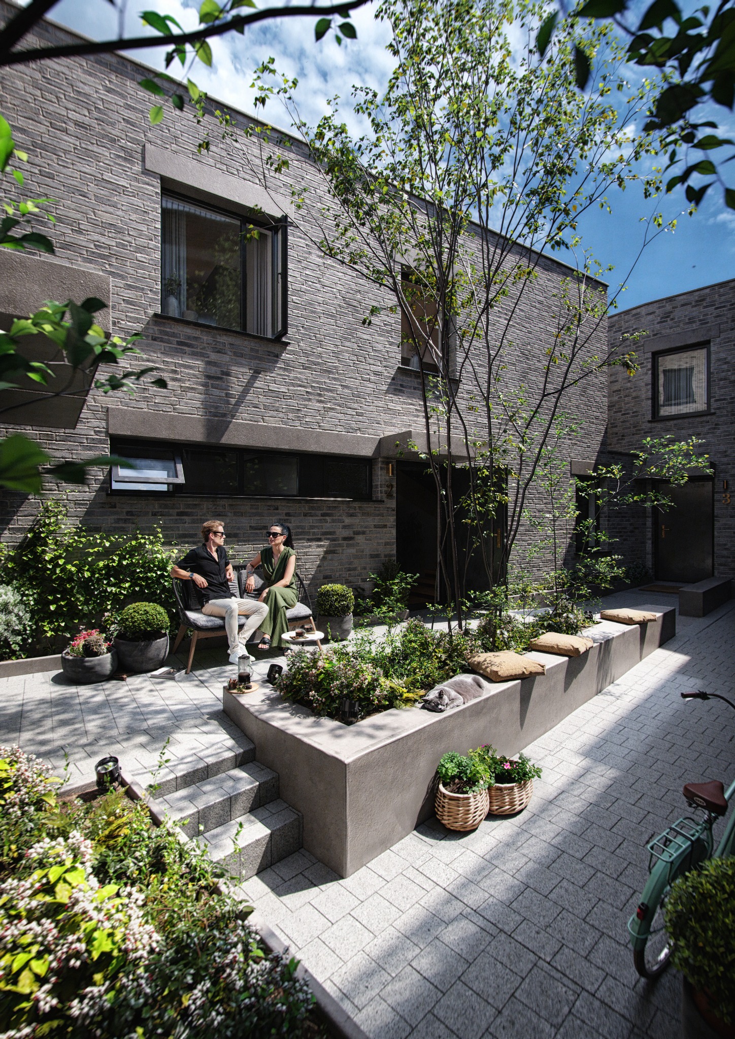 Nowoczesny dziedziniec z szarej cegły, gdzie para siedzi na ławce wśród zieleni. Kot śpi na murku, rower oparty o ścianę. Architektura minimalistyczna, rośliny w donicach.