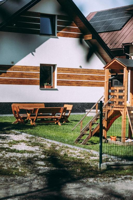 Drewniany stół z ławkami i plac zabaw przed domem z panelami słonecznymi. Dom z białymi ścianami i drewnianymi akcentami. Ujęcie z dołu, trawnik.