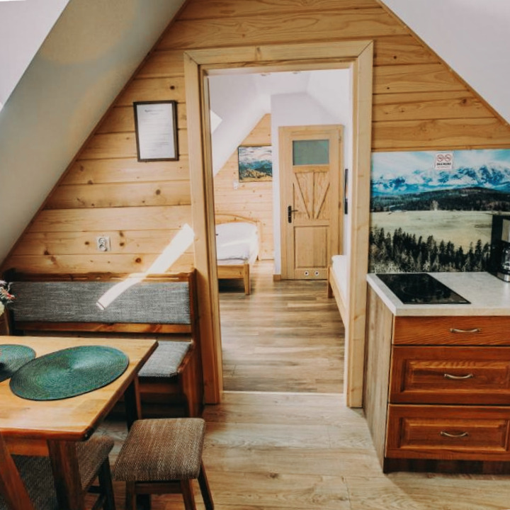 Drewniane wnętrze apartamentu na poddaszu z aneksem kuchennym, stołem i widokiem na sypialnię przez otwarte drzwi. Ukośne ściany, naturalne światło.