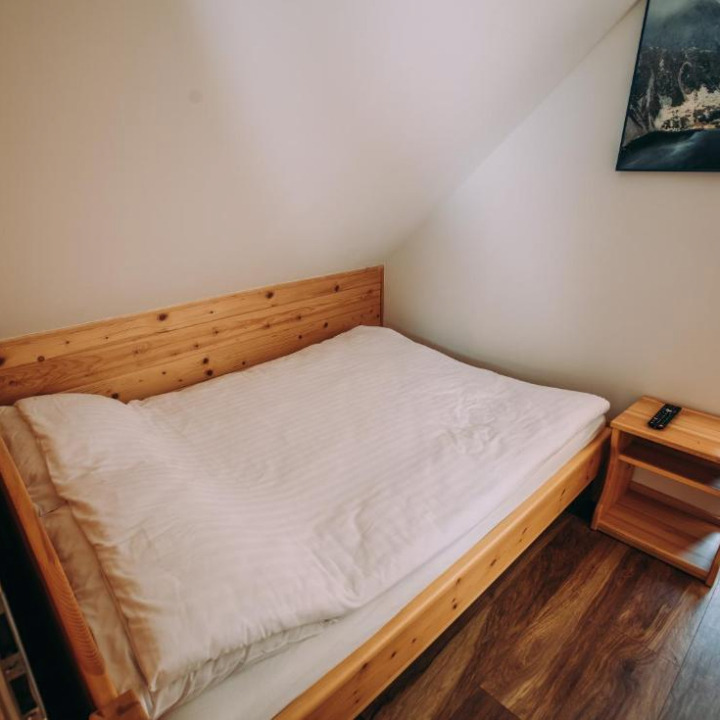 Drewniane łóżko pojedyncze z białą pościelą w pokoju na poddaszu. Obok szafka nocna z pilotem. Na ścianie obraz z motywem górskim. Ujęcie z dołu.