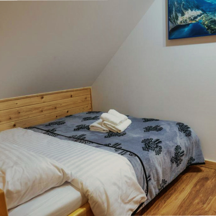 Przytulny pokój na poddaszu z drewnianym łóżkiem, wzorzystą narzutą i stosem białych ręczników. Na ścianie wisi obraz z górskim krajobrazem.