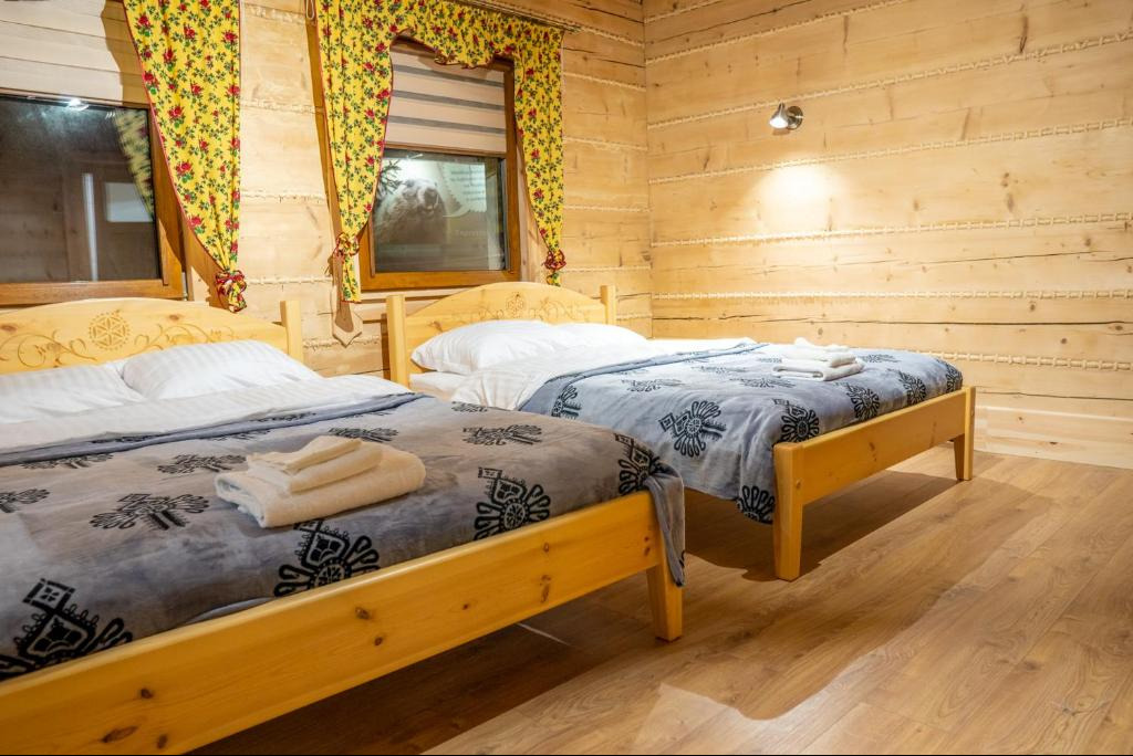 Pokój w stylu góralskim z dwoma łóżkami pojedynczymi, drewnianymi ścianami i podłogą. Okno z żółtymi zasłonami w kwiaty. Przytulne wnętrze.
