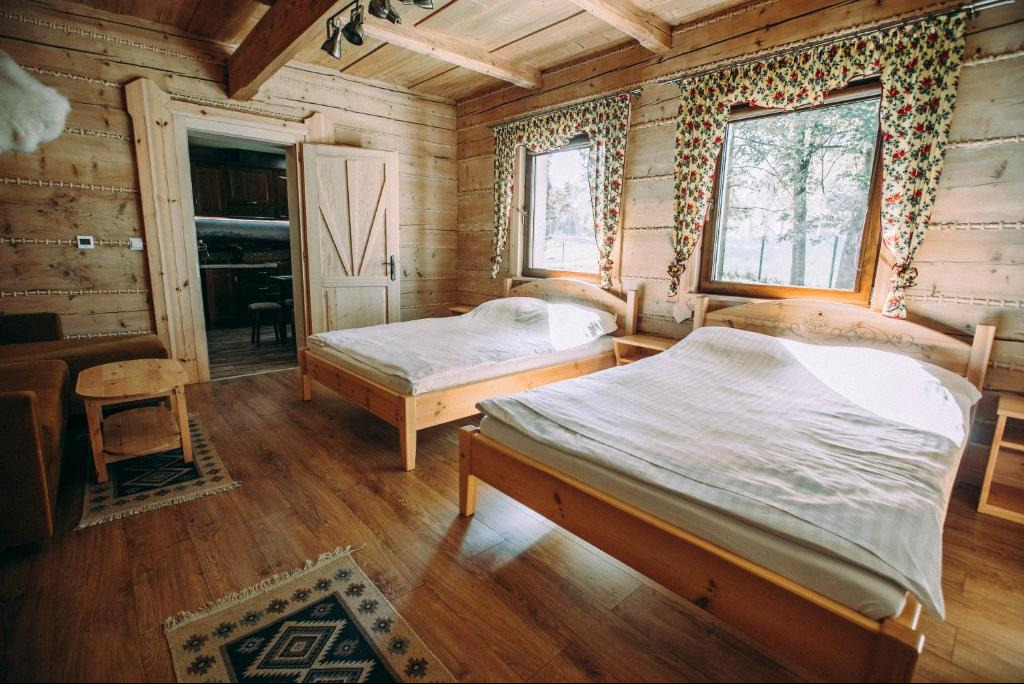 Drewniany pokój gościnny z dwoma łóżkami, oknami z kwiecistymi zasłonami i widokiem na zewnątrz. W tle widoczny aneks kuchenny.