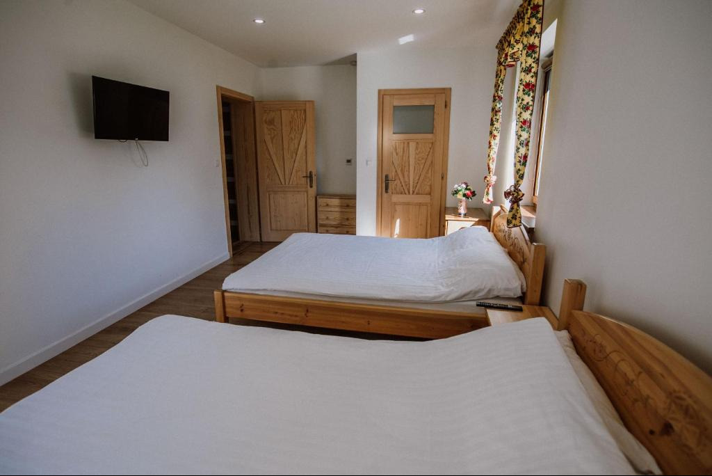 Jasny pokój z dwoma drewnianymi łóżkami, telewizorem na ścianie i kwiatami przy oknie. Widoczne drewniane drzwi z dekoracyjnymi wzorami.