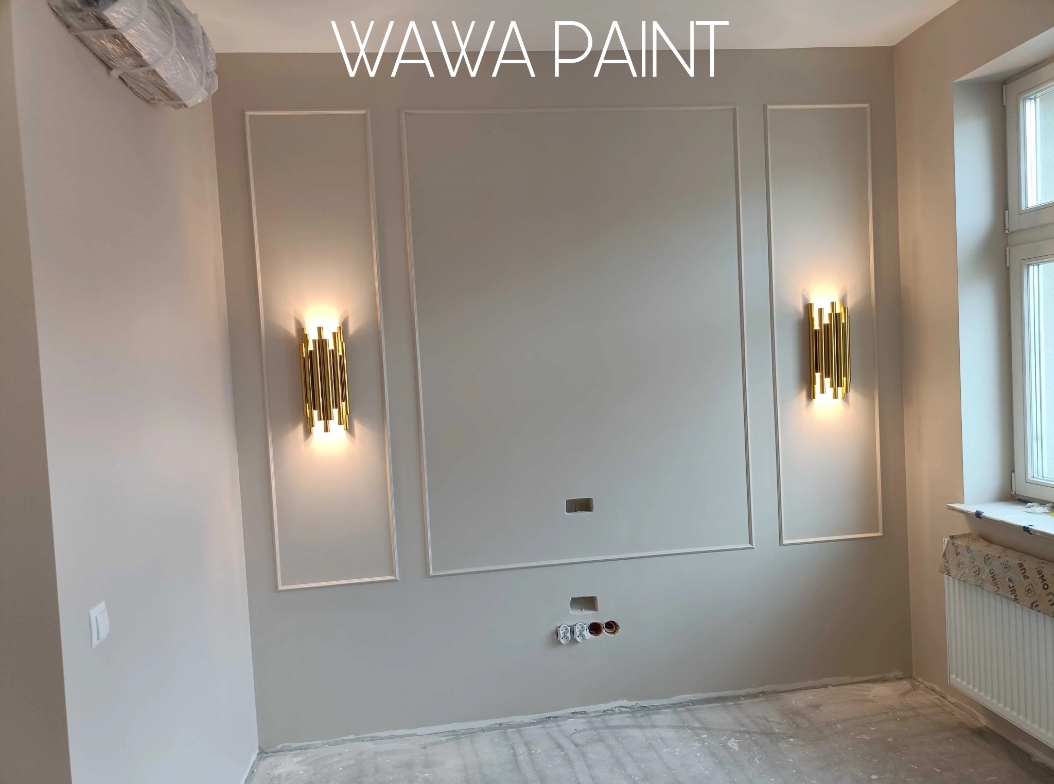 Ściana po malowaniu w odcieniach beżu z dekoracyjnymi listwami i złotymi kinkietami. Widoczne gniazdka elektryczne i zabezpieczony grzejnik.