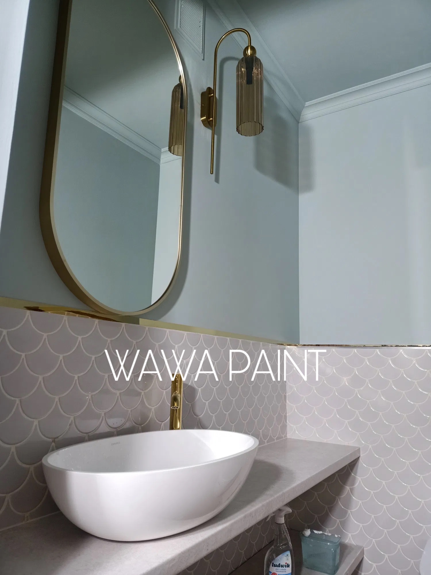 Elegancka łazienka po remoncie: biała umywalka nablatowa, złota armatura, lustro w złotej ramie i ściana w pastelowych odcieniach z mozaiką w kształcie rybiej łuski.