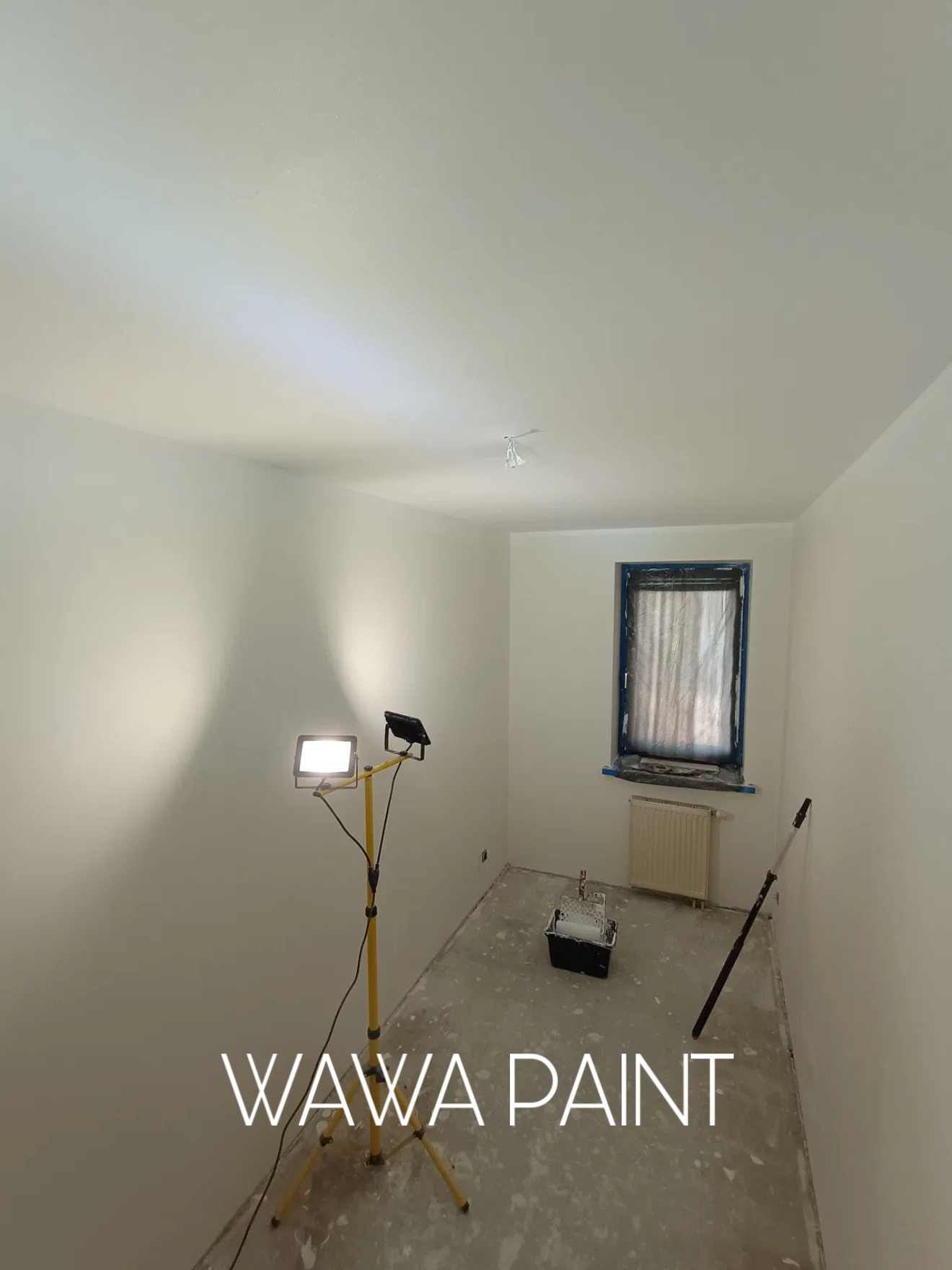 Białe, świeżo pomalowane wnętrze pokoju z oknem zabezpieczonym folią. Na podłodze widoczne ślady farby, a w rogu stoi lampa oświetlająca ścianę.
