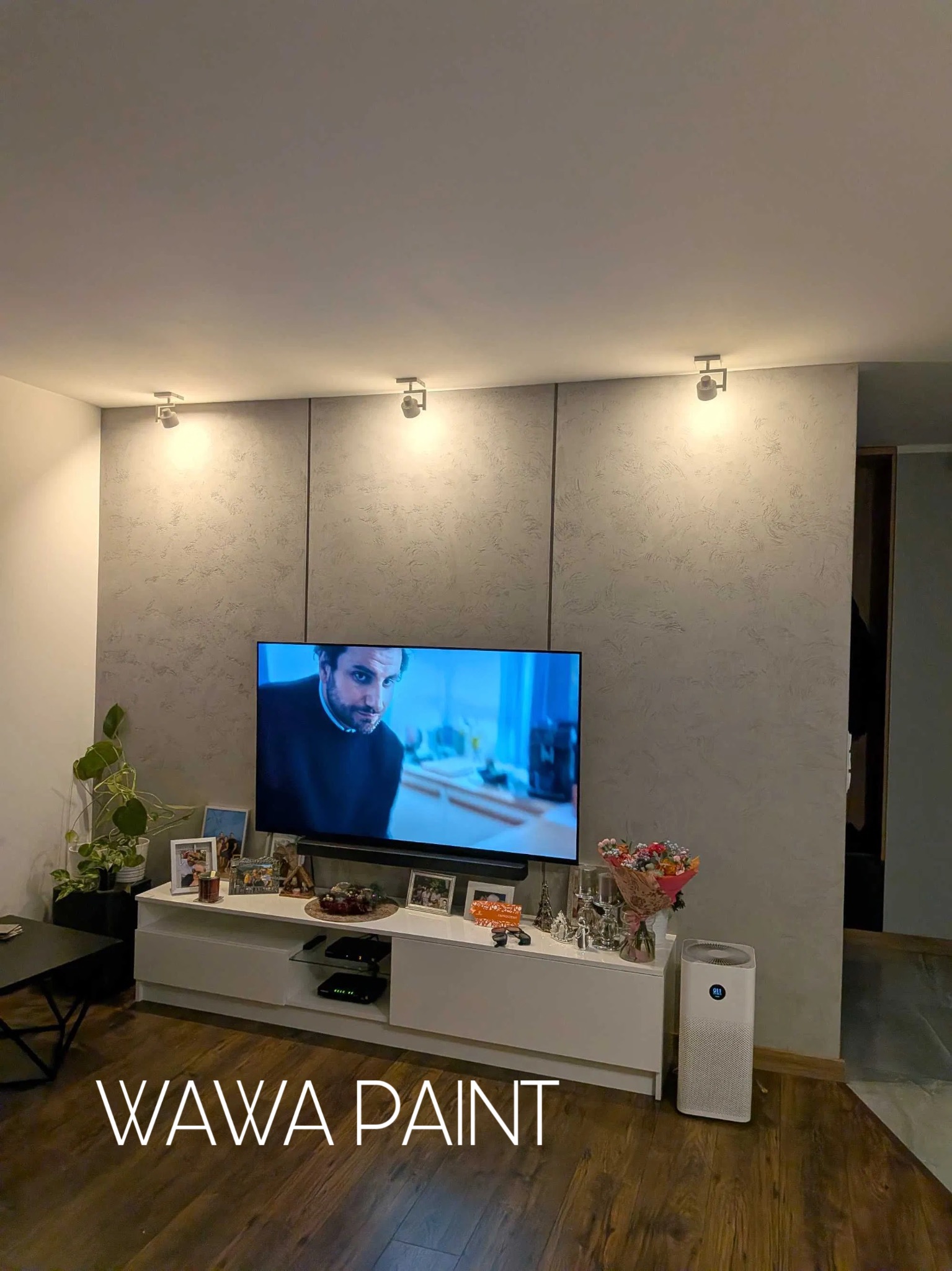 Ściana w salonie po dekoracyjnym malowaniu z efektem betonu, oświetlona trzema punktowymi lampami. Na ścianie wisi telewizor, pod nim biała szafka RTV.
