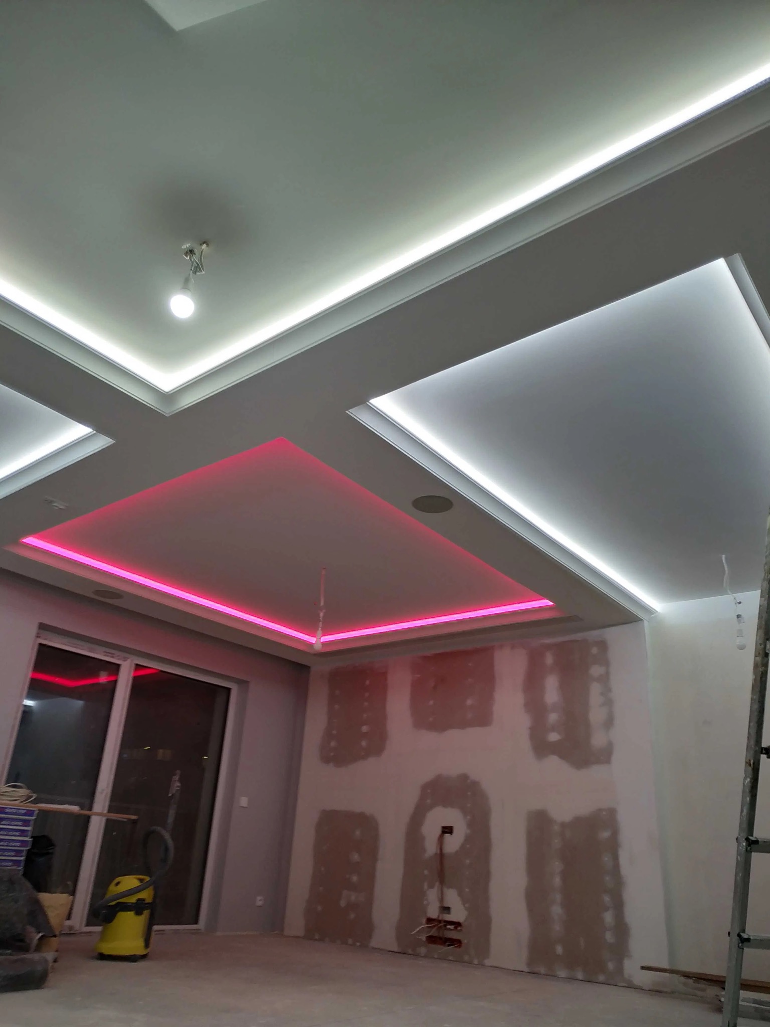 Sufit podwieszany z oświetleniem LED: białe listwy i różowe kwadraty. Ściany w trakcie szpachlowania, widoczna drabina i odkurzacz. Nowoczesne wykończenie wnętrza.