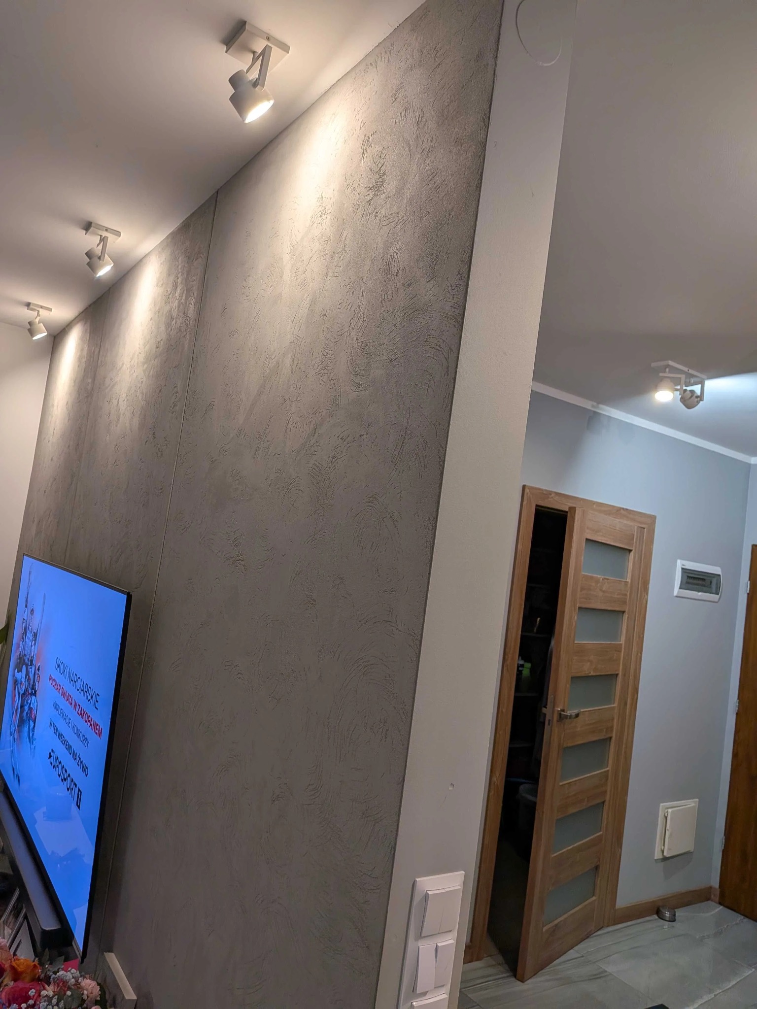 Ściana w salonie z dekoracyjnym szarym tynkiem strukturalnym, oświetlona reflektorami sufitowymi. W tle widoczne drzwi i fragment drugiego pomieszczenia.