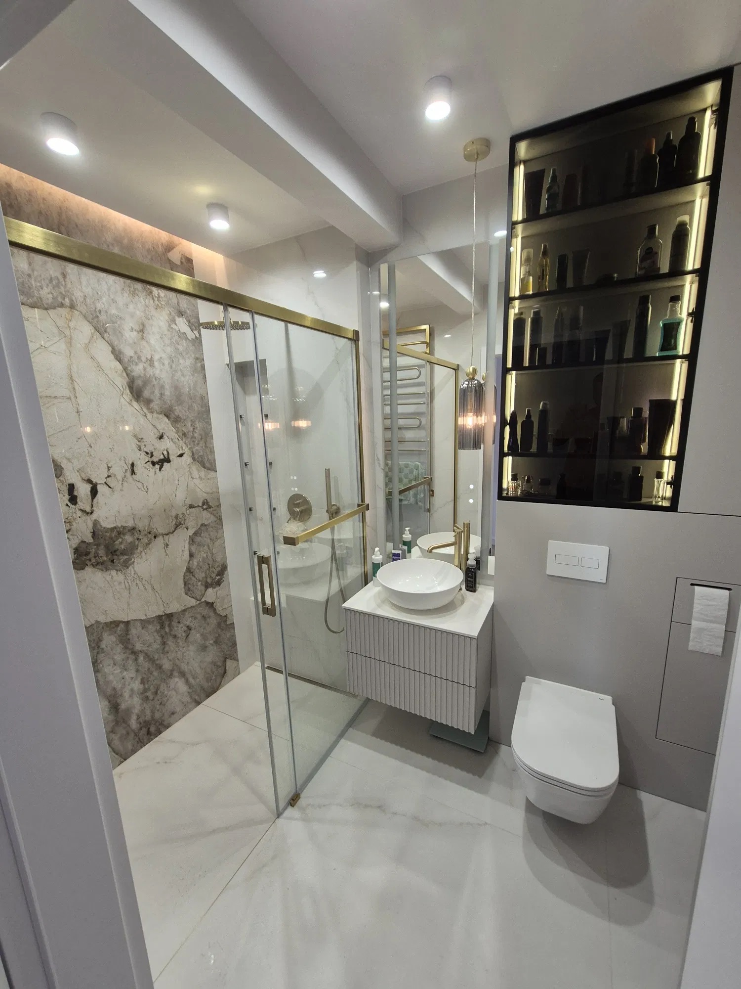 Elegancka łazienka po remoncie: kabina prysznicowa ze złotymi detalami, marmurowe ściany, biała umywalka nablatowa i toaleta. Nowoczesne oświetlenie i szafka z kosmetykami.