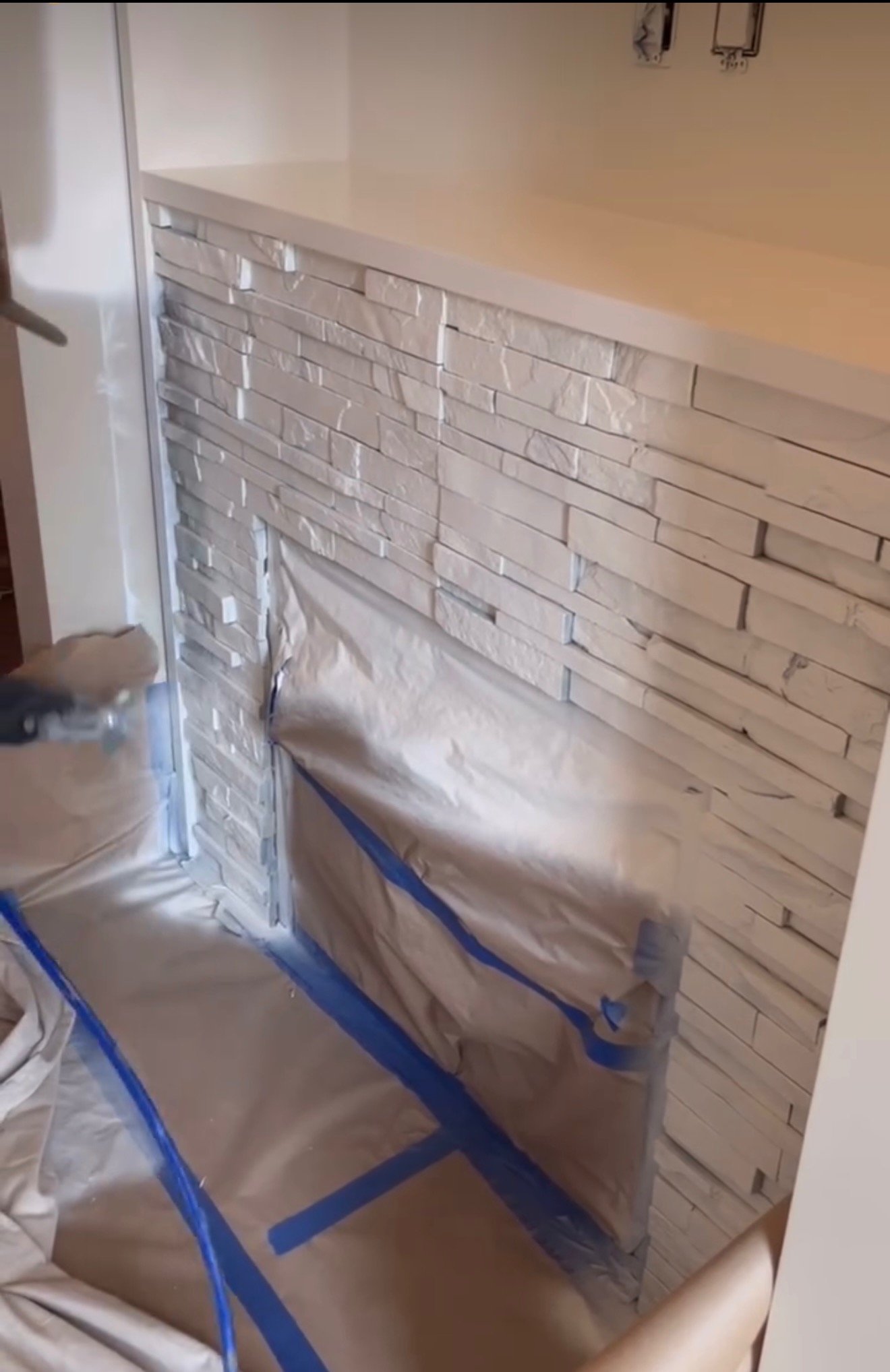 Białe kamienie dekoracyjne malowane natryskowo, zabezpieczone folią i taśmą malarską. Widoczne gniazdka elektryczne. Detal prac wykończeniowych.