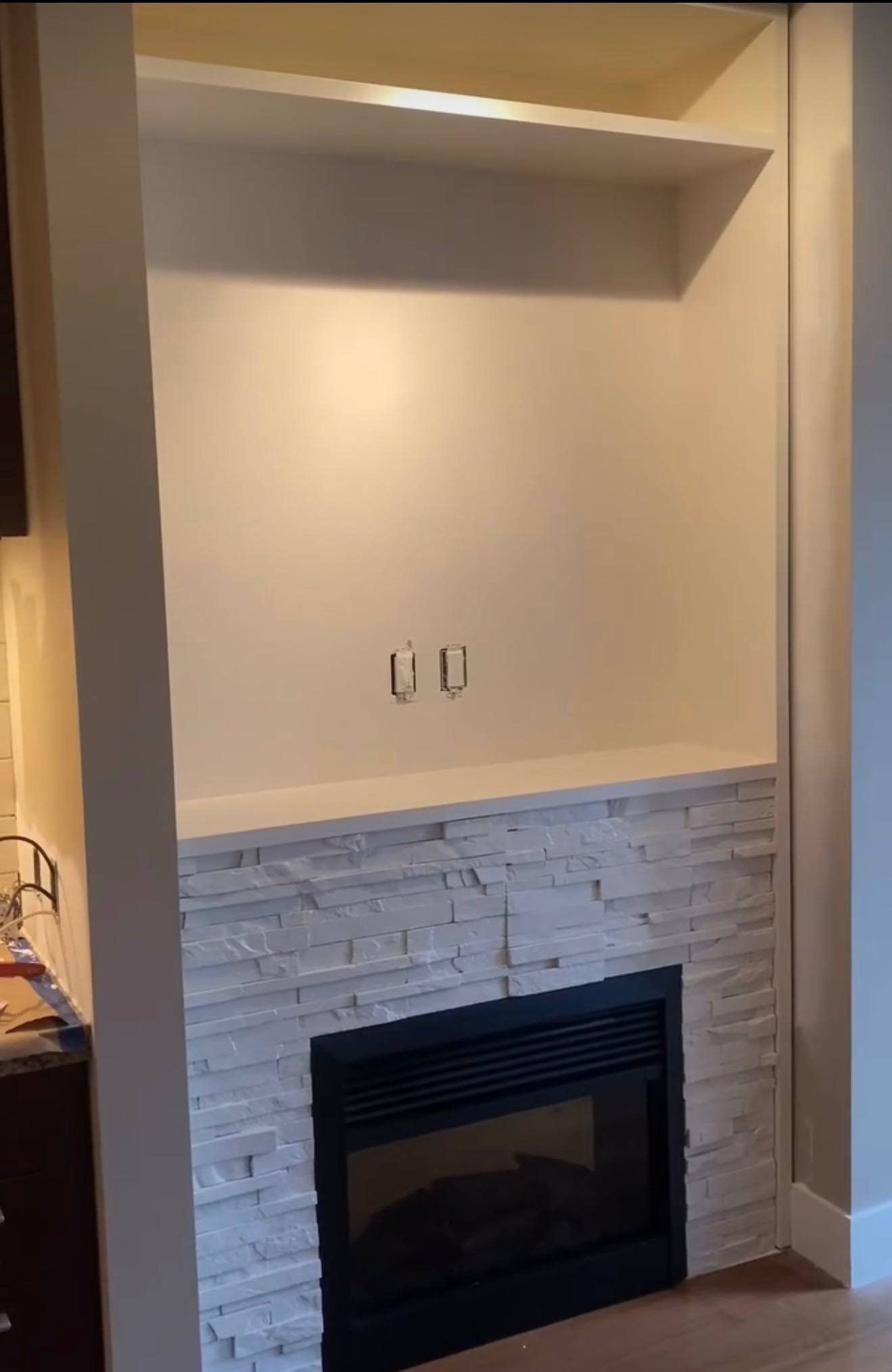 Narożny kominek z obudową z białego kamienia dekoracyjnego i wnęką na telewizor. Widoczne gniazdka elektryczne. Minimalistyczny design.