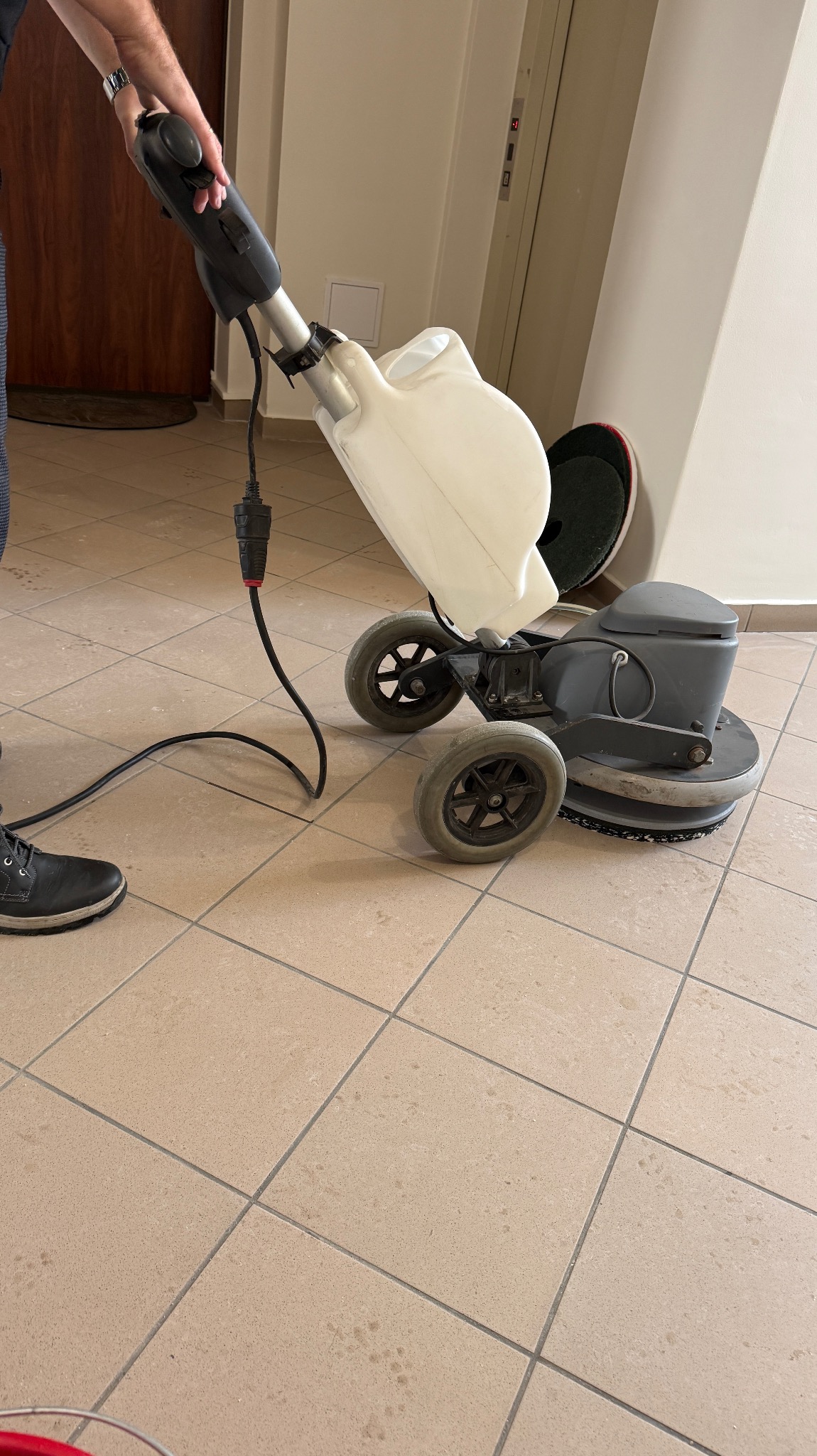 Maszyna do czyszczenia podłóg w trakcie pracy na kafelkach. Widoczny operator i kabel zasilający. W tle drzwi i ściana z windą. Sprzątanie biur.