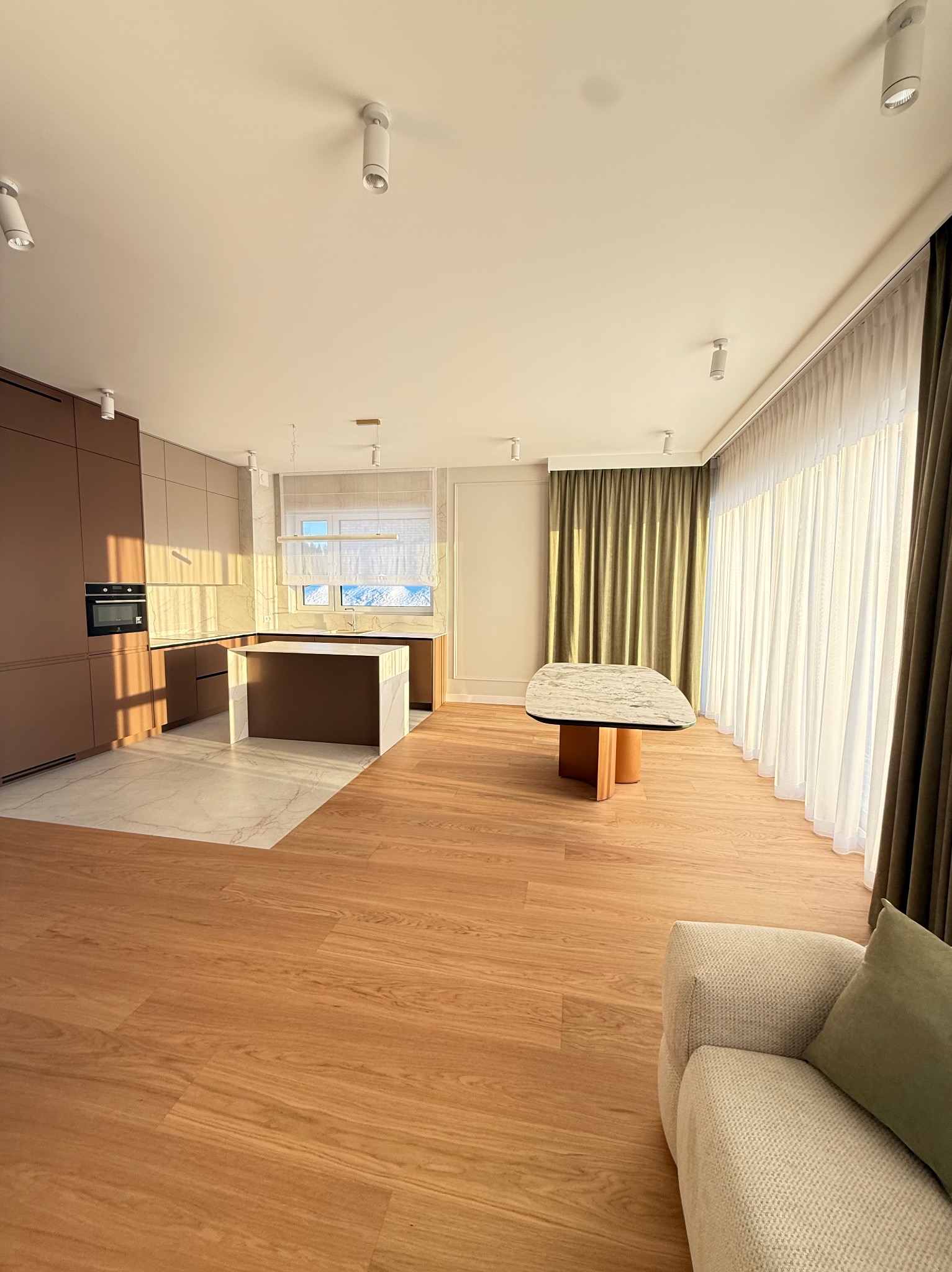 Nowoczesne wnętrze: brązowa zabudowa kuchenna z wyspą, marmurowy blat stołu, drewniana podłoga i jasne zasłony w słonecznym salonie.