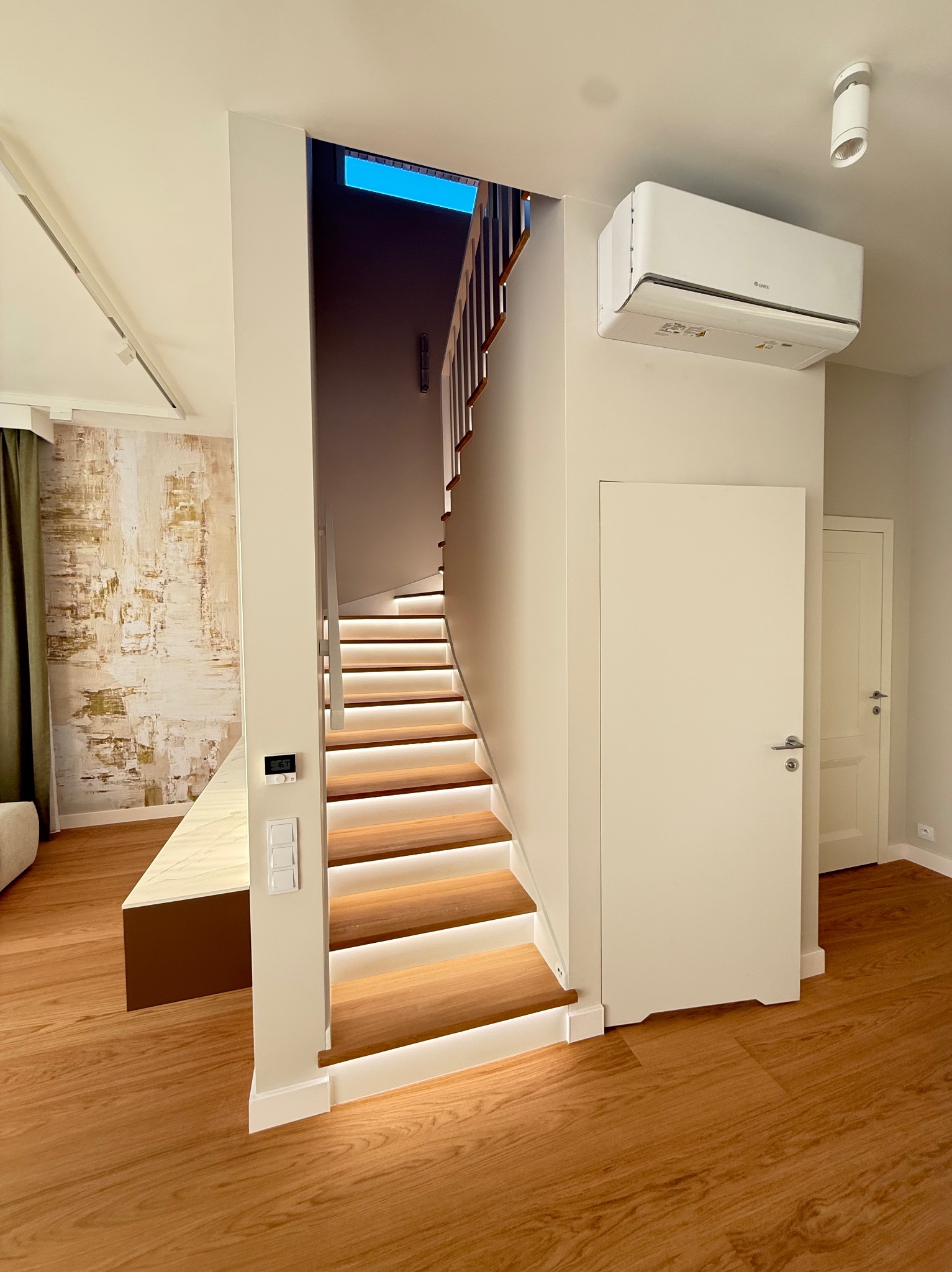 Nowoczesne schody z podświetleniem LED, drewniane stopnie, białe ściany, klimatyzator i parkiet. Widok na fragment pokoju z abstrakcyjną tapetą.
