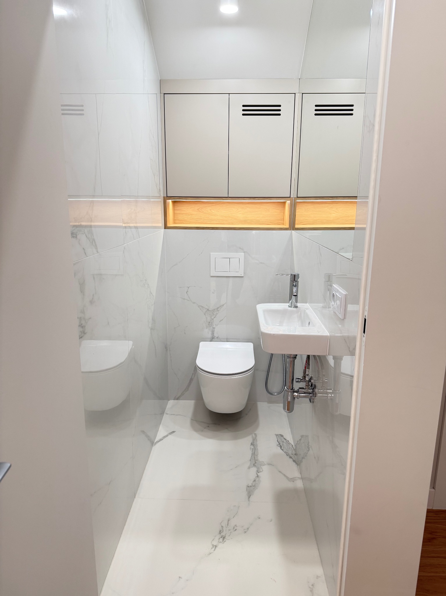 Nowoczesna, biała łazienka z marmurowymi płytkami w Pruszkowie. Umywalka z chromowaną baterią, podwieszana toaleta i szafki z oświetleniem LED.