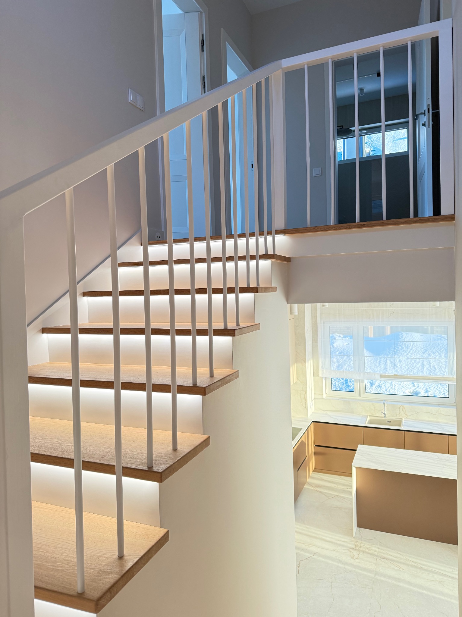 Nowoczesne schody z drewnianymi stopniami i białymi barierkami, podświetlone ledami. W tle widoczna kuchnia z oknem i śniegiem za nim.