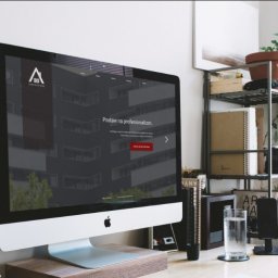 4K for Clients - Ekran iMac prezentuje stronę internetową z minimalistycznym logo i hasłem 'Postaw na profesjonalizm', na biurku z roślinami i elementami dekoracyjnymi.