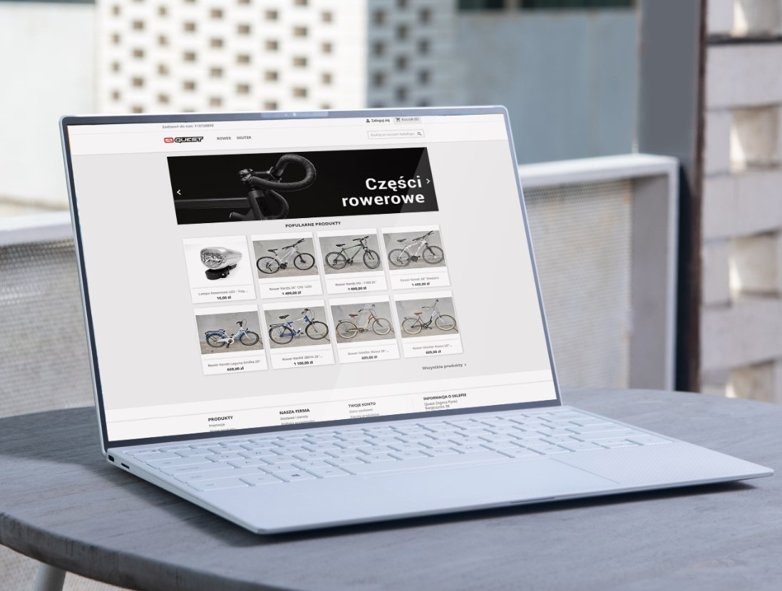 Laptop wyświetla stronę internetową sklepu rowerowego z ofertą części i rowerów. Widoczny minimalistyczny design laptopa na tle jasnego otoczenia.