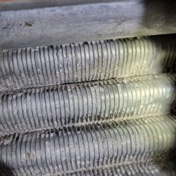 P.H.U. ARISTO - Żebrowany wymiennik ciepła pieca gazowego, pokryty białym osadem, widoczny z bliska pod kątem. Element instalacji grzewczej w trakcie przeglądu.