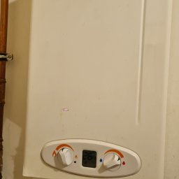 P.H.U. ARISTO - Biały, wiszący gazowy podgrzewacz wody Ferroli z widocznymi przyłączami gazowymi i wodnymi. Na obudowie naklejka informacyjna i panel sterowania z wyświetlaczem.