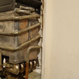 P.H.U. ARISTO - Zdemontowany, stary piec gazowy z widocznymi śladami korozji i zabrudzeń, umieszczony na tle ściany. Widoczne rury, zawory i elementy instalacji.