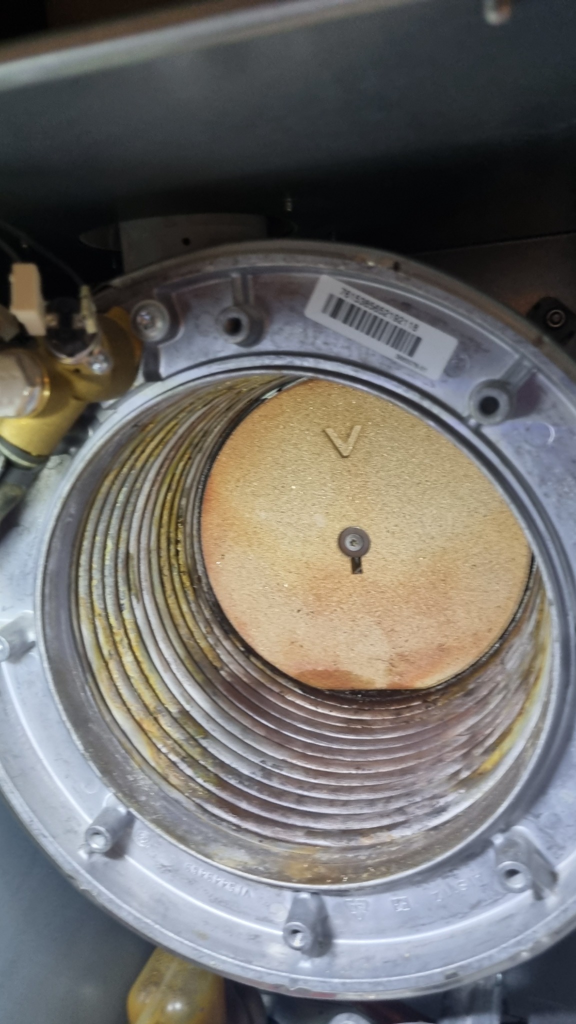 Wnętrze pieca gazowego z widocznym palnikiem i śladami użytkowania. Metalowa obudowa z widocznymi śrubami i numerem seryjnym. Palnik z symbolem 'V' i centralnym otworem.