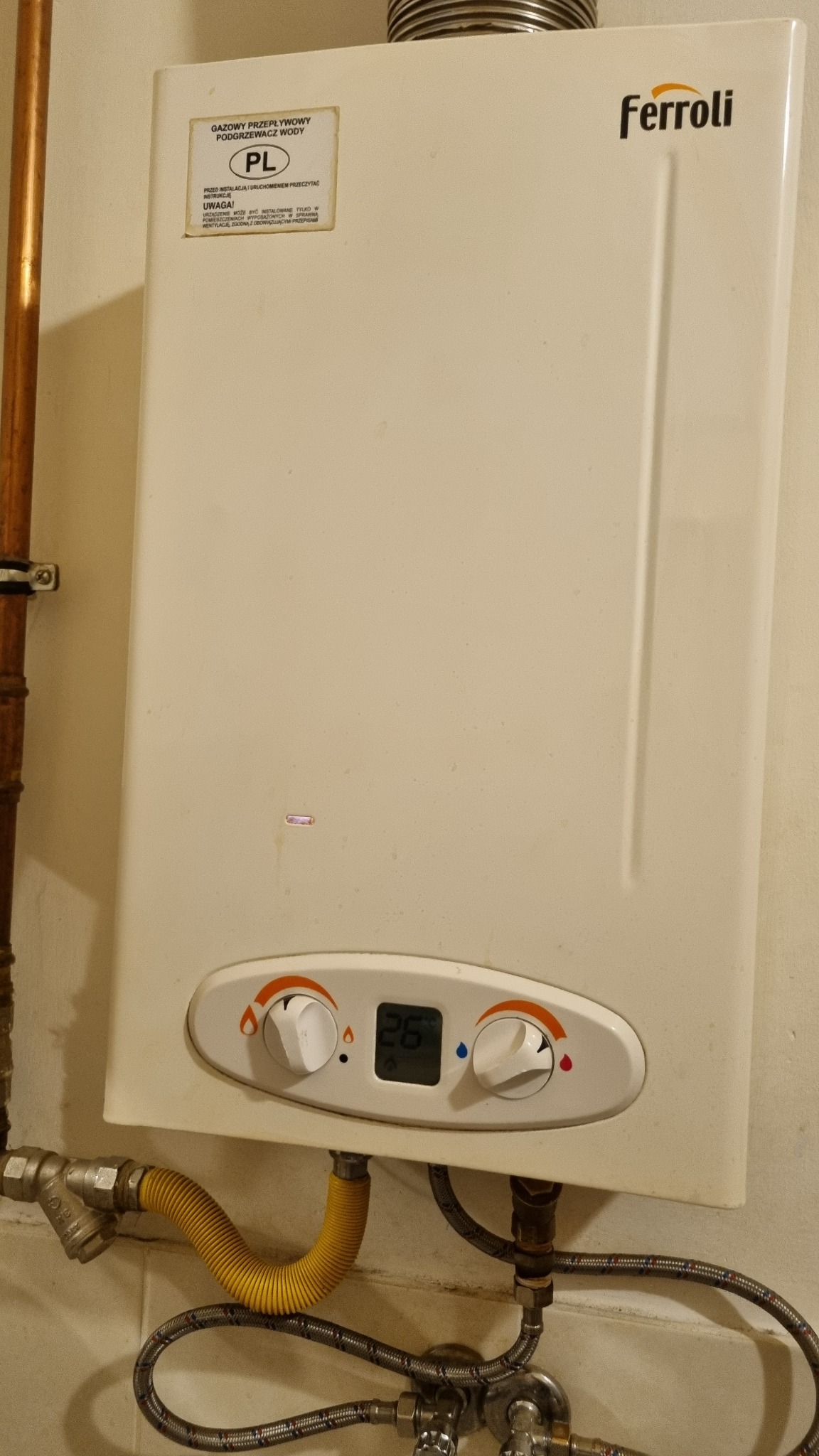 Biały, wiszący gazowy podgrzewacz wody Ferroli z widocznymi przyłączami gazowymi i wodnymi. Na obudowie naklejka informacyjna i panel sterowania z wyświetlaczem.