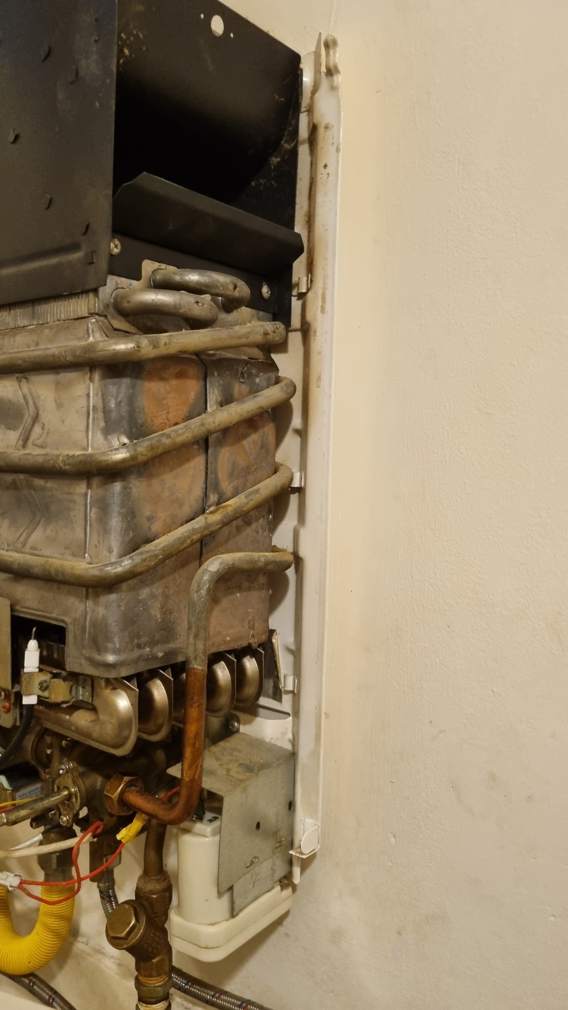 Zdemontowany, stary piec gazowy z widocznymi śladami korozji i zabrudzeń, umieszczony na tle ściany. Widoczne rury, zawory i elementy instalacji.