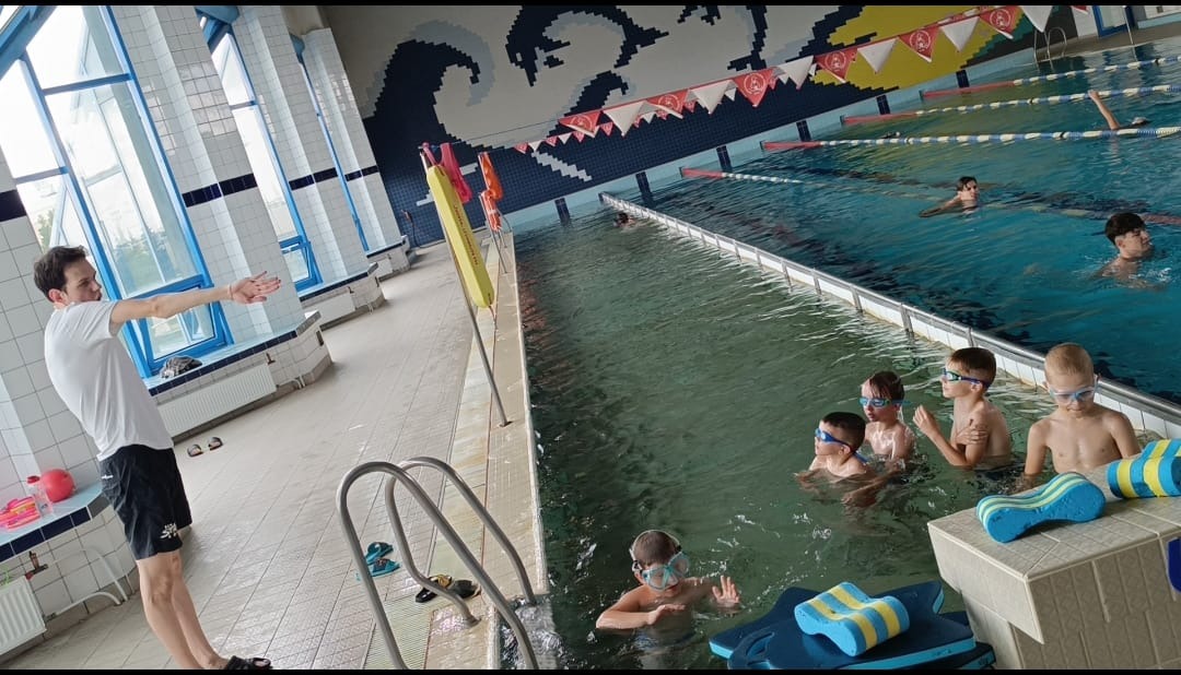 Instruktor z wyciągniętą ręką uczy grupę dzieci pływania w basenie. W tle widać inne osoby w wodzie i dekoracje na ścianach.