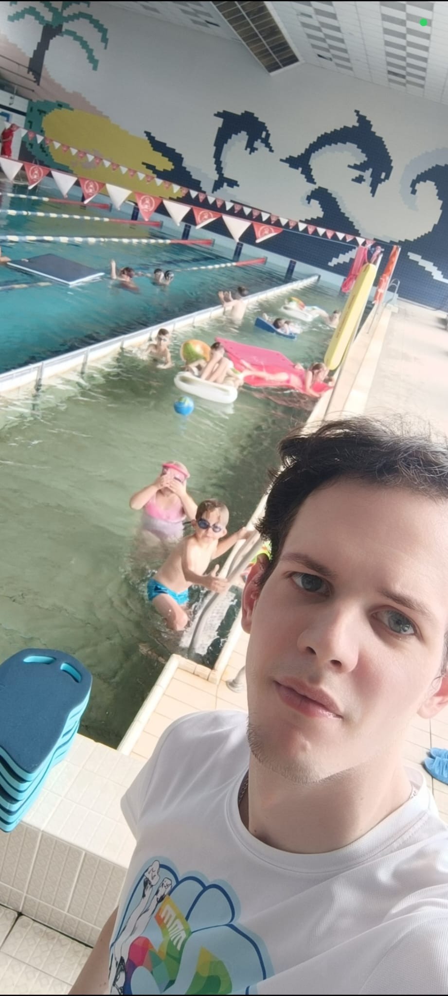 Instruktor z dziećmi w basenie podczas zajęć nauki pływania. W tle widoczne dekoracje z motywami morskimi i palmami. Uczestnicy korzystają z akcesoriów do pływania.