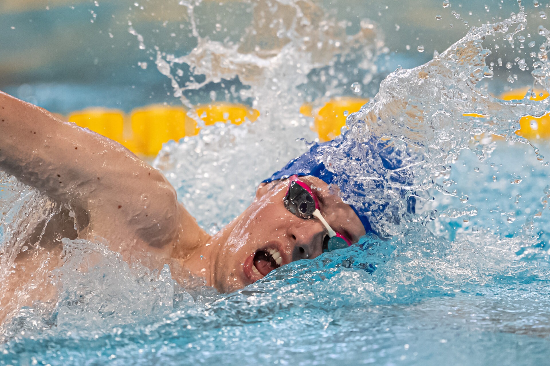Pływak w trakcie dynamicznego kraula, woda rozpryskuje się wokół głowy w niebieskim czepku i okularach, widoczne żółte pasy torów basenowych.