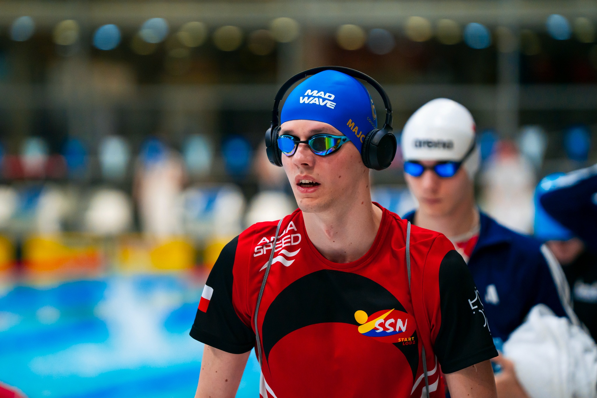 Pływak w czepku Mad Wave i słuchawkach na basenie, w okularach pływackich, w czerwono-czarnej koszulce z logo SSN Start Łódź, w tle woda i inni pływacy.
