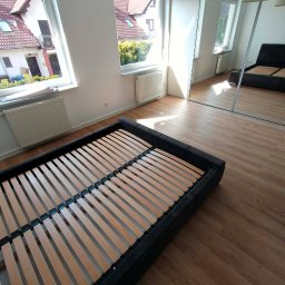 Karol - Jasne wnętrze z drewnianą podłogą i częściowo złożonym łóżkiem. Widok na okno z budynkami na zewnątrz. W tle lustrzana szafa odbijająca fragment pokoju.