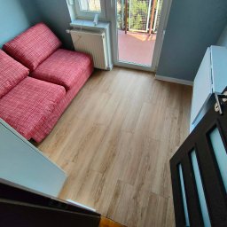 Karol - Jasne panele podłogowe w pokoju z wyjściem na balkon, widoczne z perspektywy otwartych drzwi. Czerwona sofa i grzejnik przy ścianie. Ujęcie z góry.