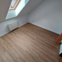 Karol - Jasne panele podłogowe w pokoju na poddaszu z oknem dachowym i grzejnikiem. Widoczny fragment drewnianej belki konstrukcyjnej.