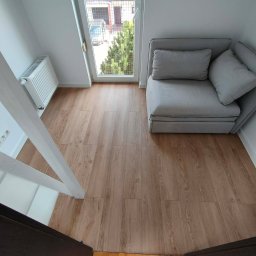 Karol - Jasne panele podłogowe w pokoju z białymi ścianami, widok z góry. Fragment schodów, okno z widokiem na zieleń i szara sofa w rogu.