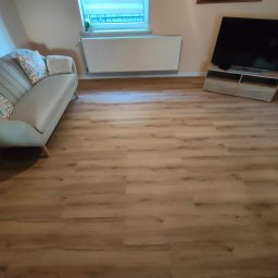 Karol - Salon z nową, jasną podłogą z paneli imitujących drewno. Widoczna sofa, telewizor i grzejnik. Ujęcie z niskiego kąta podkreśla wzór paneli.